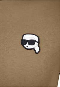 Camisa verde oliva con un parche en relieve en blanco y negro de un rostro estilizado con gafas de sol, tejido texturizado y diseño ajustado.