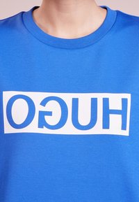 Niebieski bawełniany sweatshirt z dużą białą prostokątną grafiką z napisem "HUGO" napisanym pogrubioną, wielką literą. Gładka tekstura.