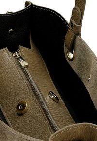 Interiér světle hnědé kožené tašky s kapsou na zip, kovovými doplňky a zapínáním na patentky. Černá podšívka kontrastuje s texturovaným světle hnědým materiálem.