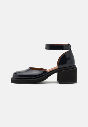 Chaussure noire en cuir verni à talon bloc, bout rond et fermeture à bride à la cheville, vue de profil sur fond blanc.