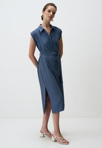 Blaues, knöpfbares Hemdkleid mit kurzen Ärmeln, tailliertem Bund, seitlichen Schlitzen und lockerer Passform, kombiniert mit weißen, sandalenähnlichen Absätzen.