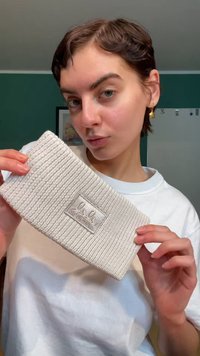 Kötött világosszürke fejpánt texturált mintával és egy fémes címkével, amelyen a márkanév szerepel, egy fehér inget viselő személy által tartva.