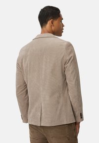 Beiger Cordjacke mit tailliertem Schnitt, Schlitz hinten und schwarzen Knopfakzenten an den Ärmeln. Glatte Textur.