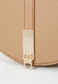 Zbliżenie beżowej skórzanej torebki, z elementami w kolorze złotym, prostokątną plakietką i akcentem logo. Widoczne szwy.