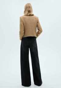 Personne aux cheveux blonds mi-longs portant une veste marron texturée courte et un pantalon noir taille haute à jambes larges, vue de dos.