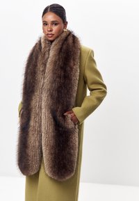 Mujer con un abrigo largo verde y una gran bufanda de piel marrón, con las manos en los bolsillos del abrigo, de pie contra un fondo blanco liso.