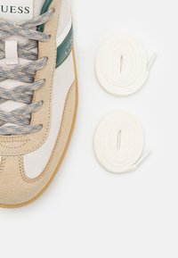 Baskets présentant une tige en daim beige et gris clair, des accents verts, des lacets plats gris, une semelle en caoutchouc gomme, et deux lacets blancs supplémentaires.