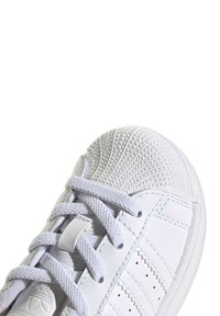White sneaker with textured rubber toe cap, smooth leather body, synthetic laces, and three side stripes for branding detail.