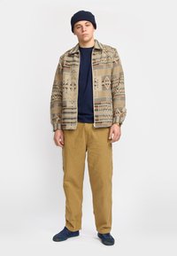 Beige Jacke mit geometrischen Mustern, darunter ein marineblaues Hemd, beige Hosen und dunkelblaue Schuhe. Das Modell trägt eine marineblaue Mütze.