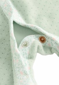 Lys grøn babyoutfit lavet af blødt stof, med en blomstret inderside, nitter i bronze og sølv, samt et struktureret ydre lag.