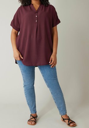 Femme portant un chemisier ample à manches courtes couleur bordeaux, un jean skinny bleu clair et des sandales plates noires à brides sur un fond uni.