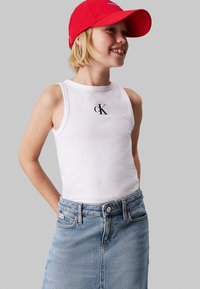 Top bianco senza maniche con il logo "CK" nero, abbinato a una gonna in denim blu chiaro. La modella indossa un cappellino rosso e sorride. Tessuto morbido e vestibilità comoda.