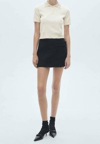 Frau, die ein beiges, kurzärmliges Polohemd, einen schwarzen Minirock mit Schnallendetails, schwarze Socken und spitze schwarze Stöckelschuhe trägt und vor einem weißen Hintergrund steht.