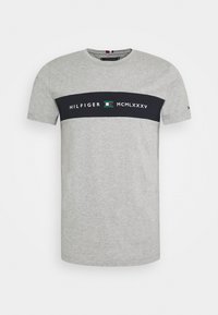 Camiseta de algodón gris con cuello redondo, que presenta una franja horizontal negra. La franja muestra "HILFIGER", "MCMLXXXV" y un pequeño emblema.