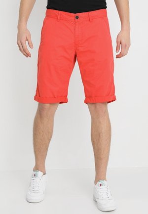 Mann trägt knallig rote hochgekrempelte Shorts, kombiniert mit weißen Reebok-Sneakern und schwarzem Oberteil vor schlichten Hintergrund.