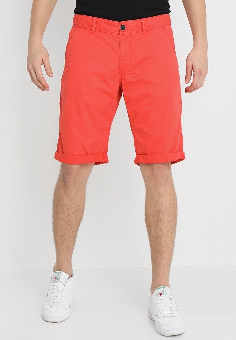 Homme portant un short rouge vif retroussé, associé à des baskets blanches Reebok et un haut noir sur un fond uni.