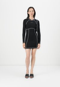 Robe noire à manches longues avec des détails en piping blanc, design ajusté et encolure ronde. Associée à des sandales plates noires.