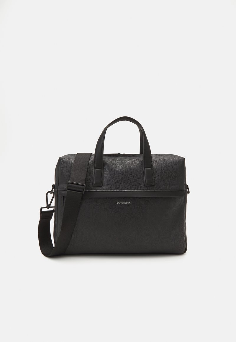 Serviette en cuir noir Calvin Klein avec double poignée, bandoulière réglable et logo à l’avant sur fond blanc uni.