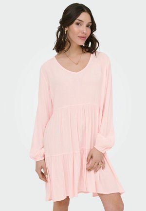 Femme portant une robe rose clair, ample, à manches longues avec une jupe à volants, debout devant un fond clair uni.