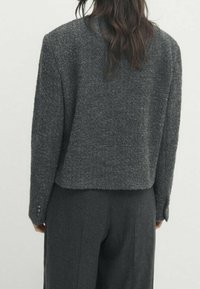 Veste grise texturée, courte, avec manches longues et poignets boutonnés, portée avec un pantalon gris foncé assorti. Design simple et moderne.