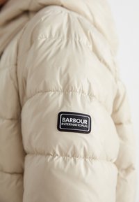 Casaco puffer bege com textura acolchoada e um logótipo preto com a inscrição "BARBOUR INTERNATIONAL" na manga esquerda.