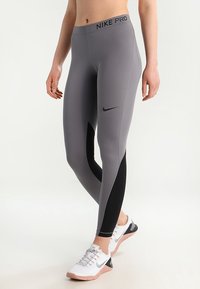 Osoba ubrana w szare i czarne legginsy Nike Pro oraz białe sneakersy Nike, stojąca z jedną nogą lekko zgiętą.
