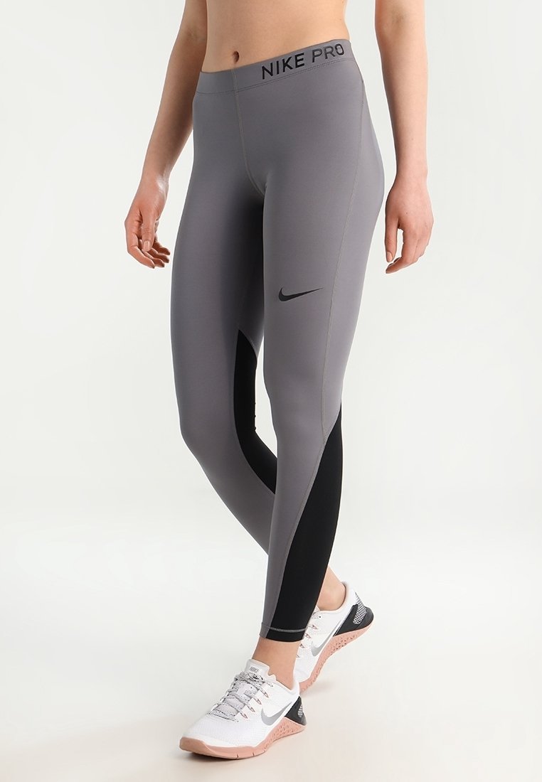 Osoba ubrana w szare i czarne legginsy Nike Pro oraz białe sneakersy Nike, stojąca z jedną nogą lekko zgiętą.