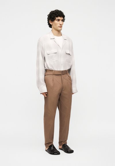 Άνδρας που φοράει ανοιχτό μπεζ πουκάμισο με μακριά μανίκια, καφέ ψηλόμεσο παντελόνι με ζώνη και μαύρα πλεκτά παπούτσια χωρίς κορδόνια, στέκεται μπροστά σε λευκό φόντο.
