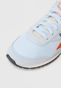 Gaiši zili sporta apavi ar teksturētu acu materiālu augšdaļu un zamša akcentiem, plakaniem šņorēm un oranžu logotipa detaļu sānos. Melna zole.