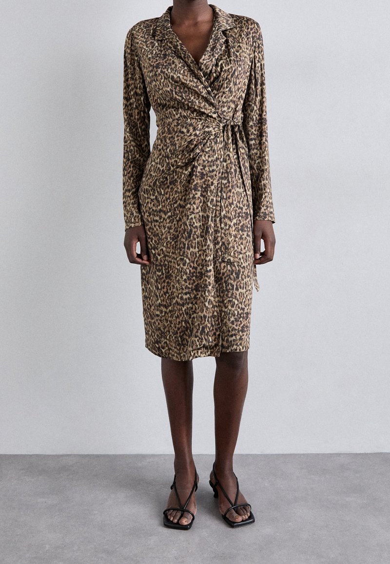 Femme portant une robe portefeuille imprimée léopard, à manches longues, longueur genoux, avec des sandales noires à brides, debout sur un sol gris face à un mur uni.