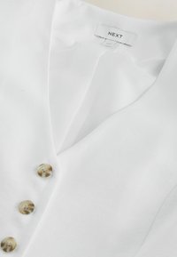 Blusa blanca con escote en V, tres botones jaspeados beige y una etiqueta de marca "NEXT" en la parte trasera del cuello.