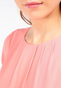 Rosa chiffongklänning med rundad halsringning och delikat plissering runt kragen, som visar en mjuk, flödande textur.