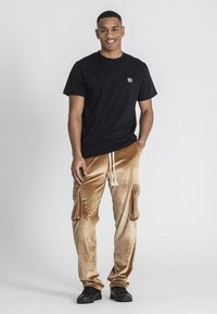 Camiseta de algodón negra y pantalones cargo de terciopelo dorado con bolsillos laterales y cordón ajustable. Modelo con zapatos negros. Fondo neutro.