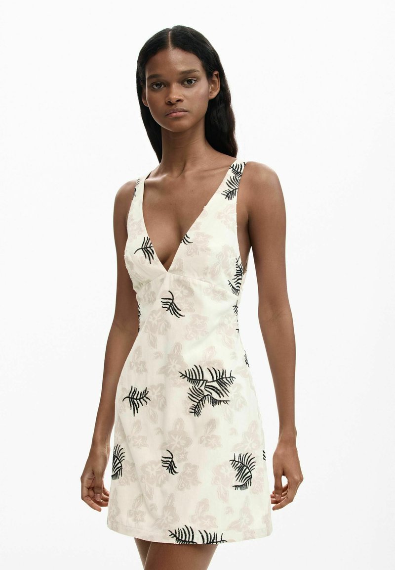 Žena u bijeloj haljini bez rukava s crnim uzorkom listova i dubokim V-izrezom, stoji na jednostavnoj bijeloj pozadini.