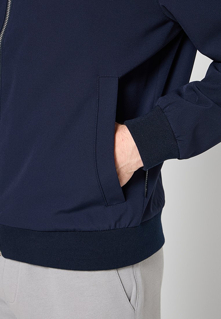 Mano en el bolsillo de una chaqueta azul marino con puño acanalado, cremallera visible, combinada con pantalones gris claro.