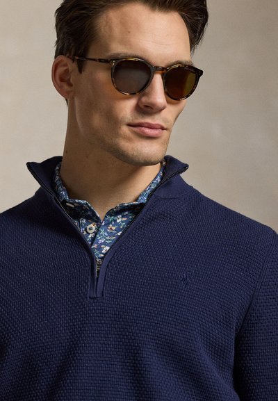 Férfi teknőcpáncél mintás napszemüveget visel, valamint egy tengerészkék, texturált cipzáros pulóvert egy kék virágmintás, gombos ing felett.