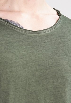 Zbliżenie na górną część klatki piersiowej osoby ubranej w zieloną koszulkę z okrągłym dekoltem, z częściowo widocznym czarno-szarym tatuażem w pobliżu obojczyka.