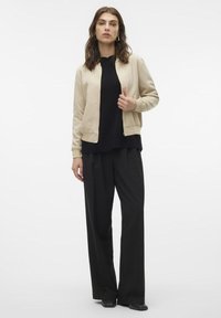 Beige Bomberjacke mit Reißverschluss und gerippten Bündchen über einem schwarzen Top, kombiniert mit locker sitzenden schwarzen Hosen. Glatte Textur und lässiges Design.