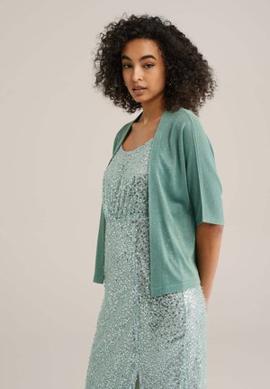 Un cardigan vert clair à manches courtes sur une robe scintillante garnie de sequins dans des tons verts assortis, présentant un ourlet droit et une fente sur le côté.