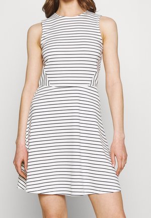 Femme portant une robe blanche sans manches avec de fines rayures horizontales noires, cintrée à la taille et évasée en une jupe courte.