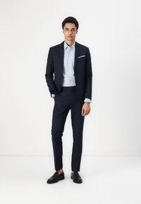 Navy suit with slim fit, single-breasted jacket, peaked lapels, and a light blue dress shirt. Black leather loafers and white backdrop.