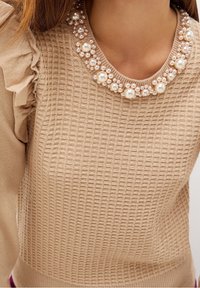 Pull en tricot beige avec un motif texturé et des détails de manches volantées, doté d'un col orné de perles composé de perles grandes et petites.