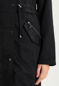 Svart jacka med dragsko i midjan, eleganta dragkedjor och tryckknappar. Tillverkad av texturerat tyg med sidodetalj i form av dragkedja.