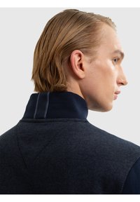 Jonge man met blond haar die een donkerblauwe trui met hoge kraag draagt, kijkt naar rechts met een neutrale uitdrukking tegen een lichtgrijze achtergrond.