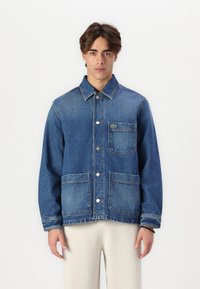Jeans jakna u plavoj boji s dugmadi sprijeda, šiljastim ovratnikom i dvije velike prednje džepove. Ima mali zeleni logo na prsima.