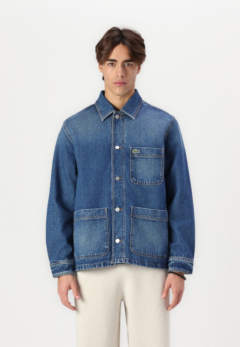 Jeans jakna u plavoj boji s dugmadi sprijeda, šiljastim ovratnikom i dvije velike prednje džepove. Ima mali zeleni logo na prsima.
