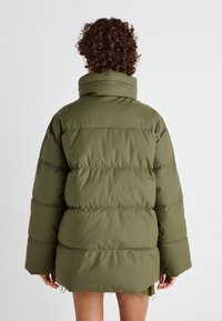 Doudoune verte olive avec une silhouette surdimensionnée, un col haut et un design matelassé horizontal. Texture douce et lisse ; manches longues.