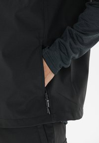 Schwarze Softshelljacke mit einer Reißverschlusstasche, darunter ein strukturiertes graues Langarmshirt. Das Design ist schlank und modern.
