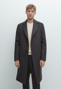 Uomo con i capelli chiari che indossa un lungo cappotto di lana grigio scuro sopra un maglione di maglia color crema, in piedi contro uno sfondo grigio chiaro uniforme.