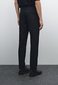 Czarne spodnie na miarę z gładkiej tkaniny, o dopasowanym kroju, z detalami tylnych kieszeni, noszone z czarnymi butami na teksturowanej podłodze.